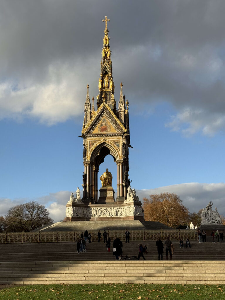 ケンジントンガーデンのアルバート記念碑を階段の下から見上げた写真。ゴシック様式の金色の壮大な記念碑が、青空と暗い雲を背景に太陽の光を浴びて輝いている。