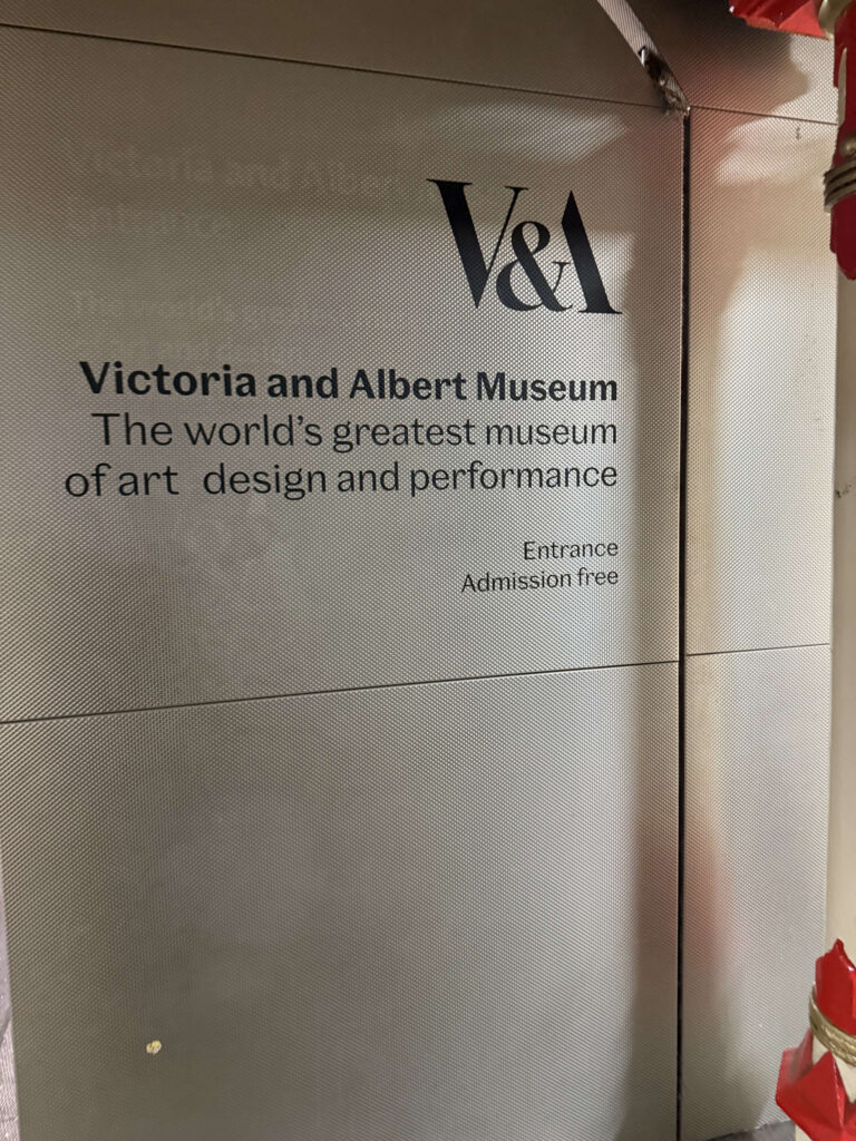 The entrance sign for the Victoria and Albert Museum on a metallic, textured wall. The black text reads 'V&A Victoria and Albert Museum The world's greatest museum of art design and performance Entrance Admission free'.