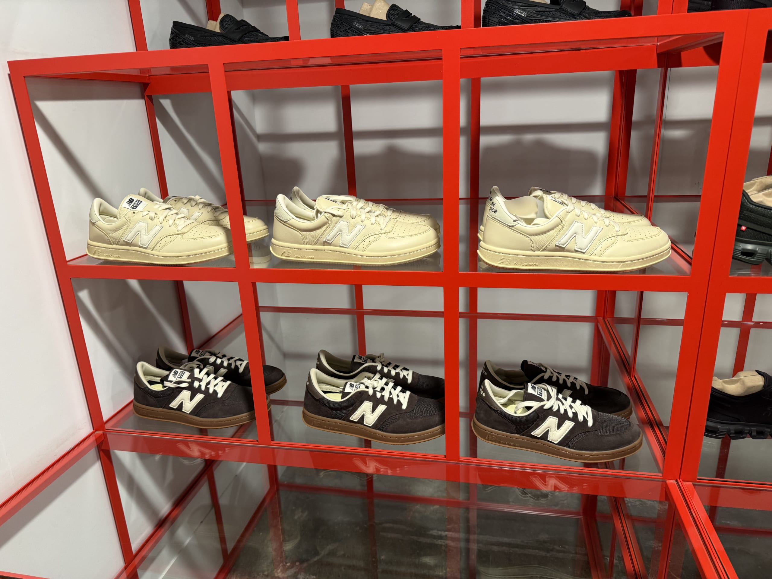 Cream and dark brown Auralee x New Balance collaboration sneakers displayed on a bright red shelving unit