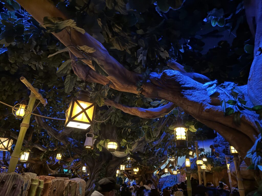 Warm glowing lanterns hanging from large artificial tree branches in a dimly lit, blue-hued indoor queue area