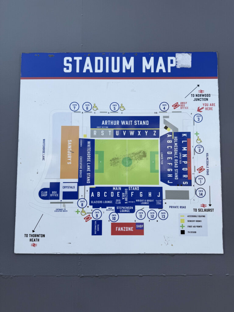A weathered outdoor sign displaying the stadium map of Selhurst Park, showing the layout of the stands, numbered entrances, and directions to nearby train stations.