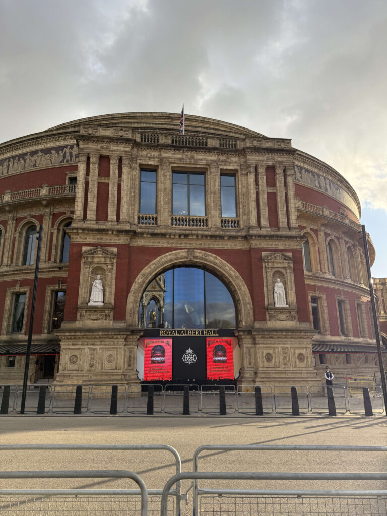 The front of the Royal Albert Hall in London against a cloudy sky. It is a grand, circular building made of red brick and stone, with a large arched window and digital screens displaying event posters at the entrance.