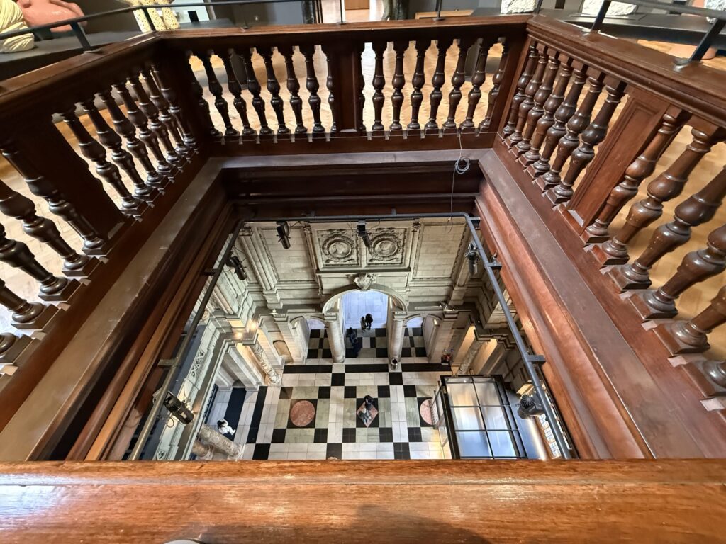 A photo looking down from an upper floor with a wooden banister into a building's atrium. Below, a black and white checkered floor spreads out, with a few people appearing small in the distance.