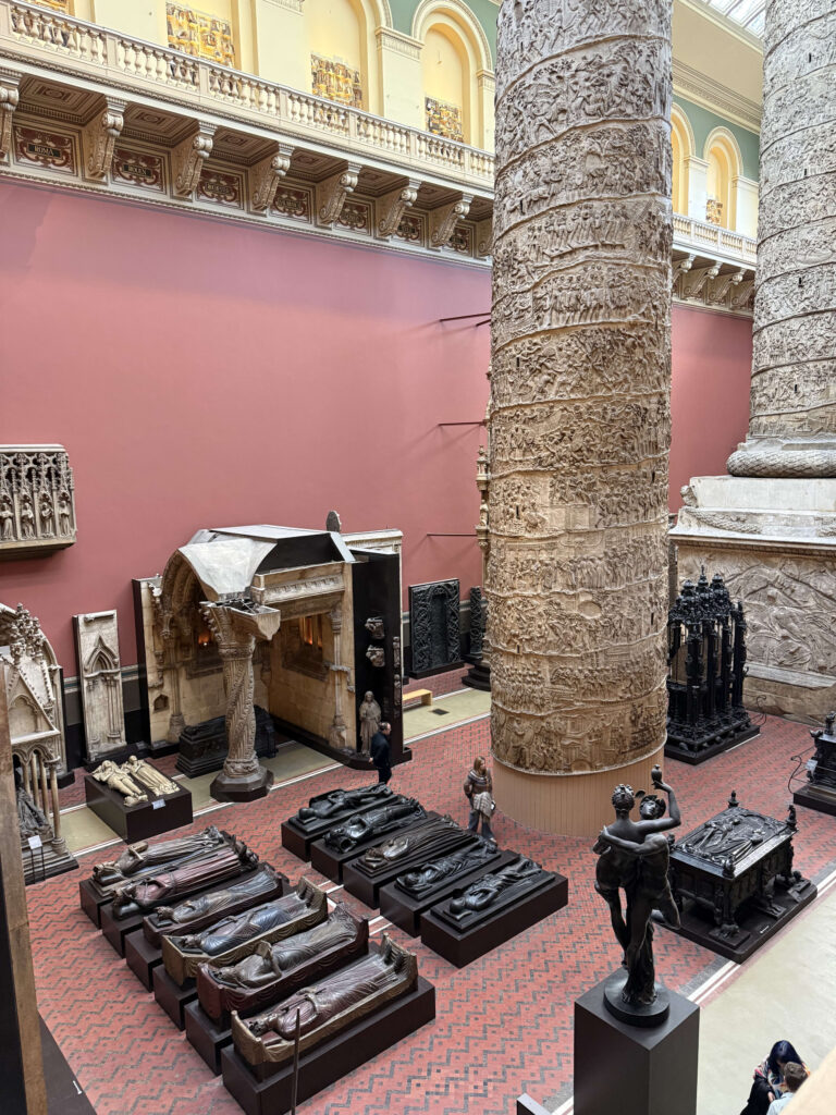 An overhead view of the Cast Courts at the Victoria & Albert Museum. Two enormous replicas of Trajan's Column tower over the gallery floor, which is filled with numerous replicas of sarcophagi and sculptures.