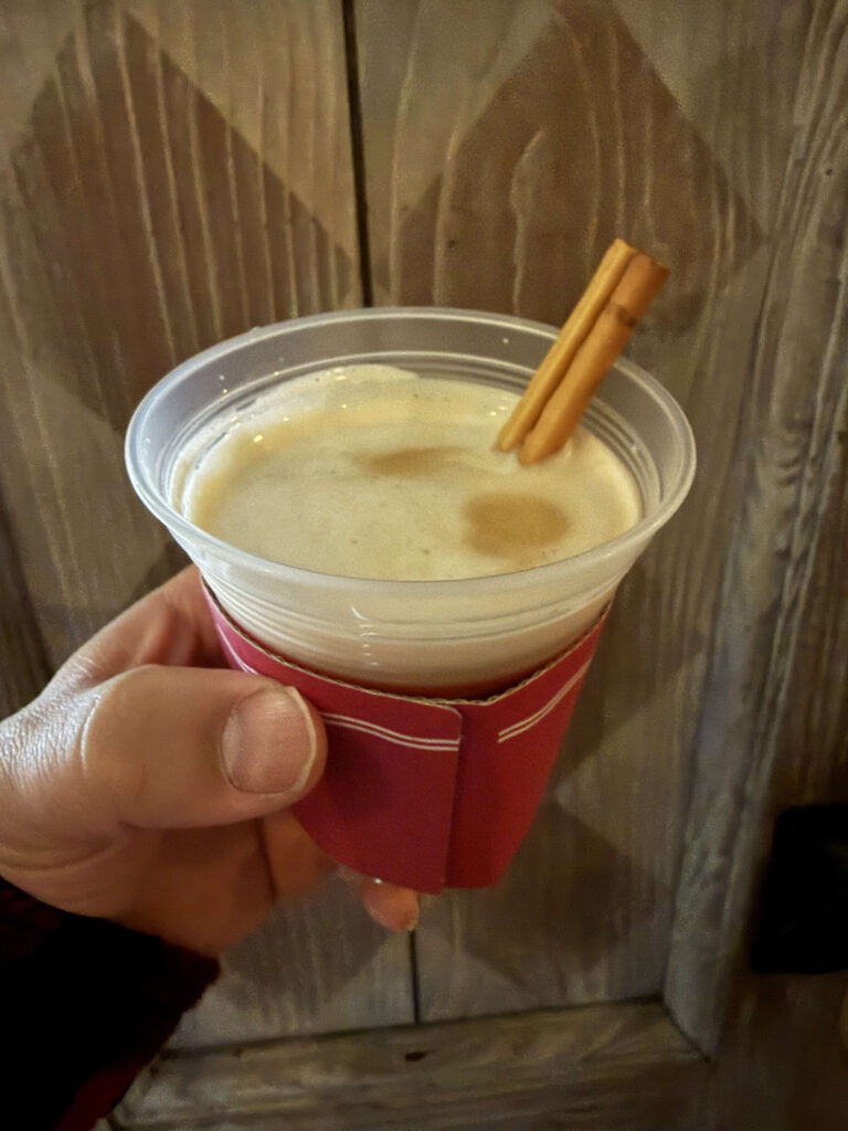 A hand holding a plastic cup with a red sleeve containing a creamy hot cocktail garnished with two cinnamon sticks