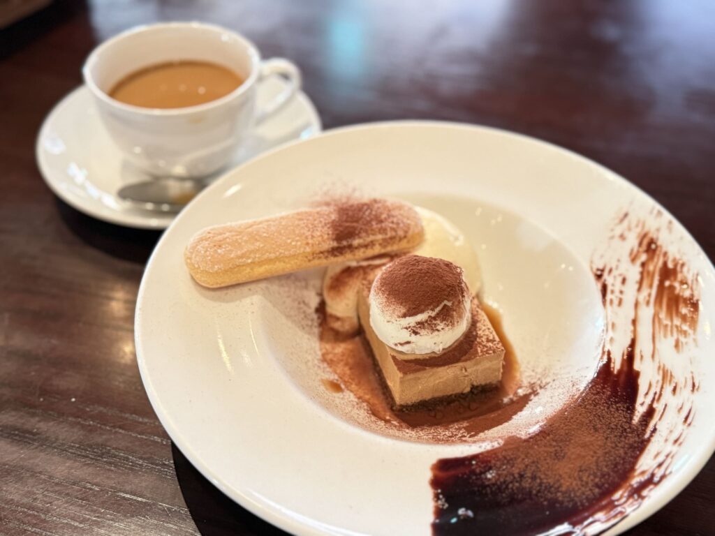 Plated tiramisu dessert with a ladyfinger biscuit and a cup of coffee in the background