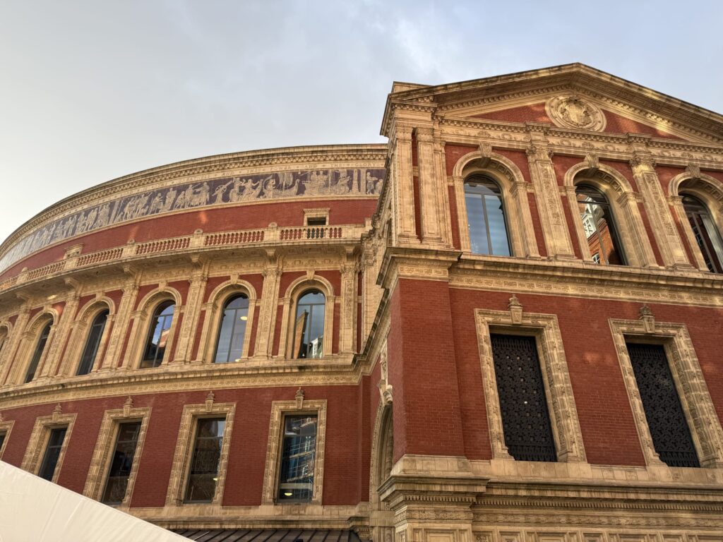 The exterior of the Royal Albert Hall against a lightly clouded sky. The magnificent building, made of red brick and beige stone, is captured from a low-angle perspective.