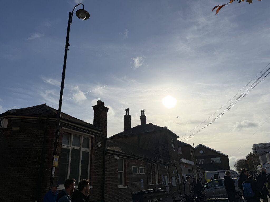 青空と輝く太陽を背景に、逆光でシルエットになったノーウッド・ジャンクション駅のレンガ造りの外観と街灯