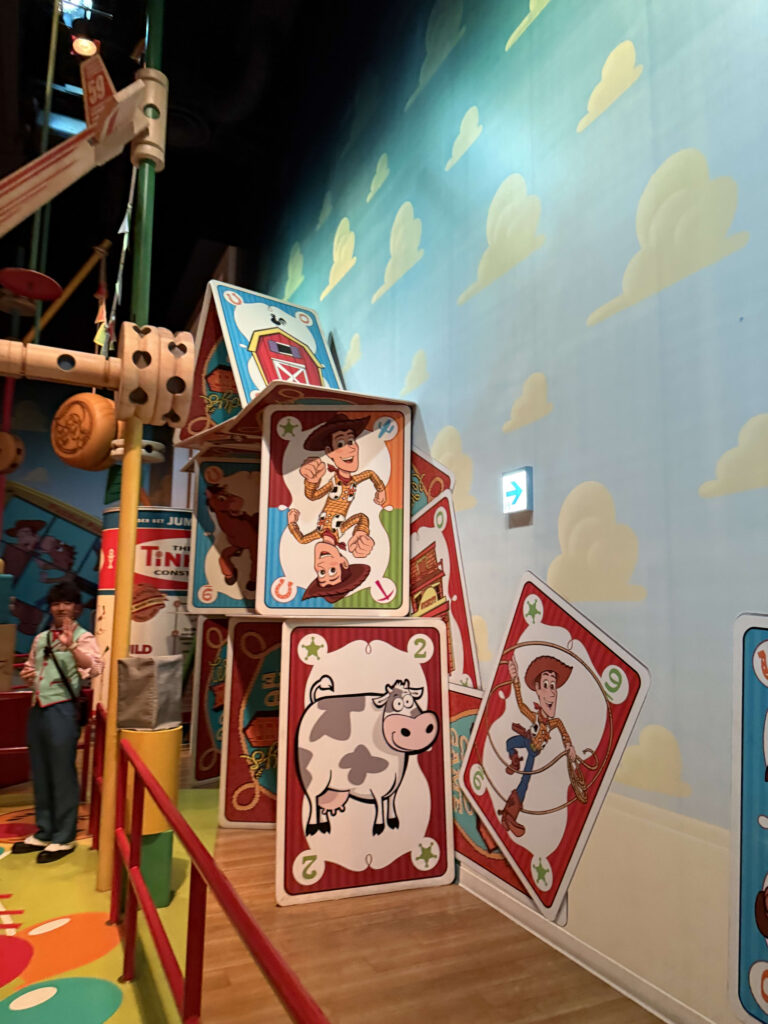 Interior of an attraction featuring giant playing cards depicting Woody from Toy Story and a wall with a blue sky and white cloud pattern