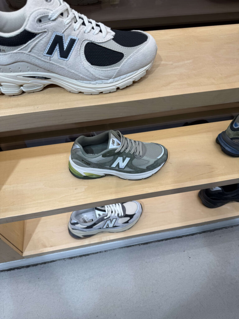 New Balance sneakers displayed on wooden shelves in a store, featuring white, olive green, and grey models.