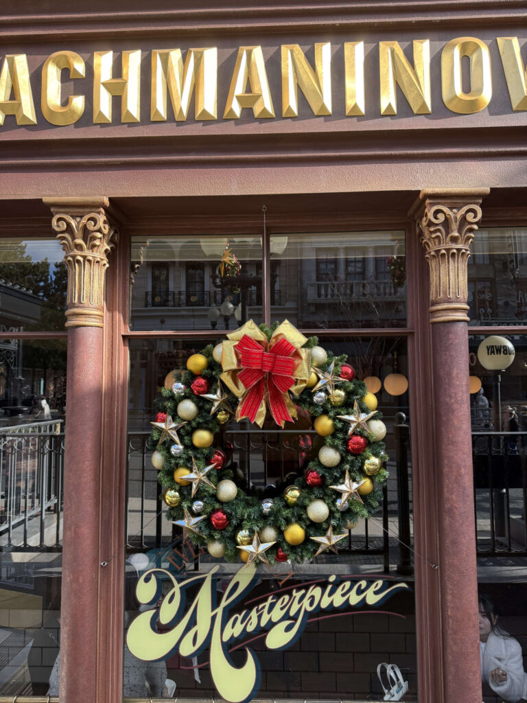 A festive Christmas wreath with a large red bow and gold stars hanging in a shop window with classical columns and a sign that reads RACHMANINOV.