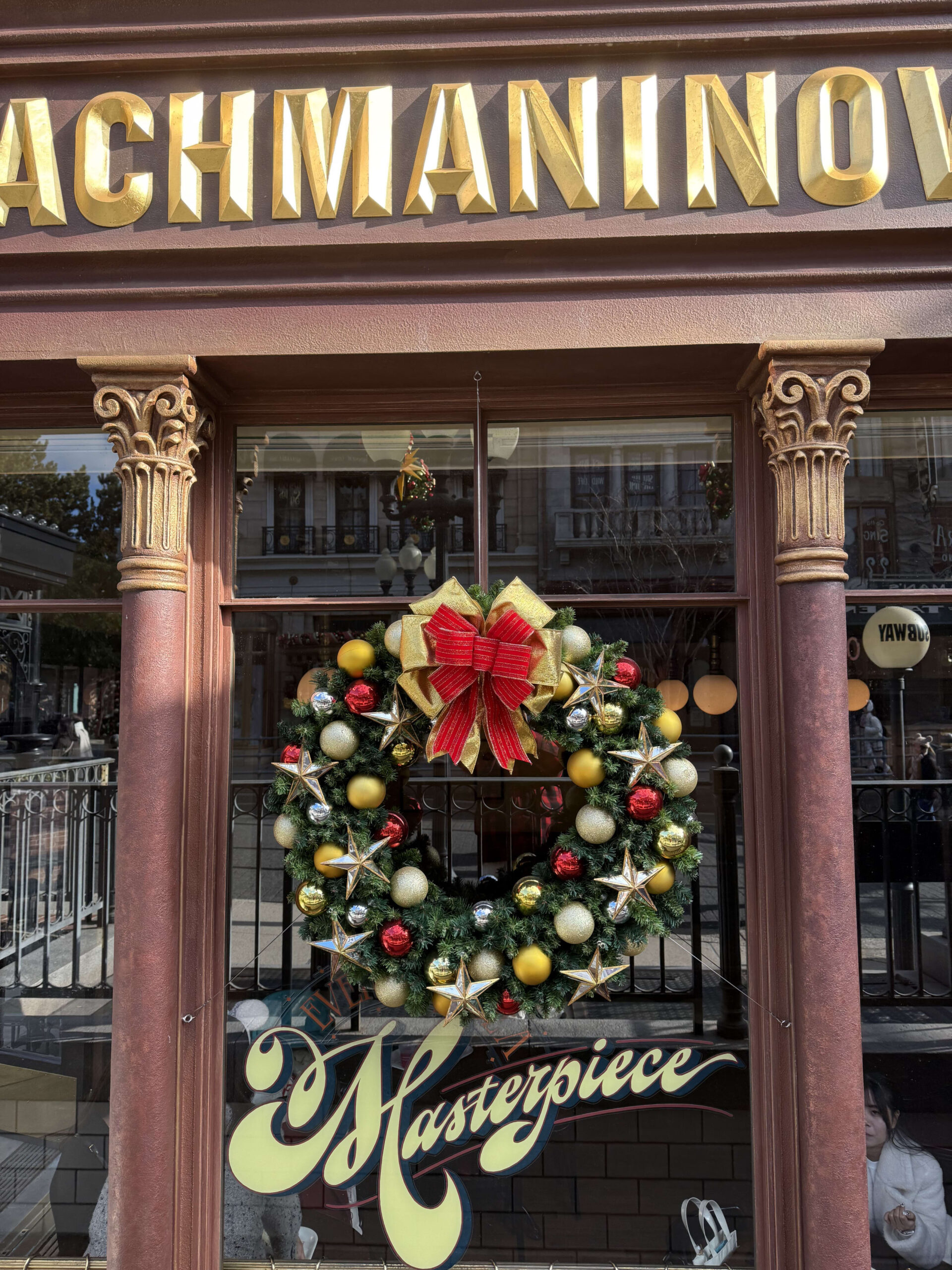 A festive Christmas wreath with a large red bow and gold stars hanging in a shop window with classical columns and a sign that reads RACHMANINOV.