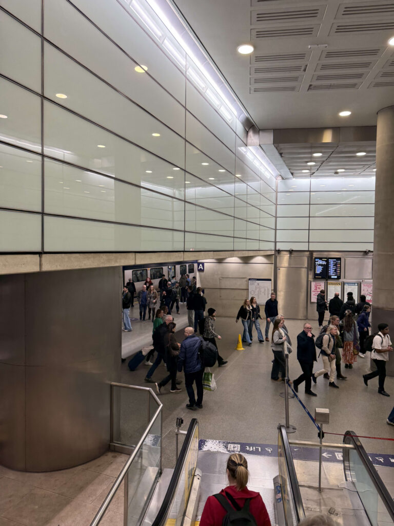 エスカレーターの上から見下ろした近代的な駅のコンコース。左側には大きな曇りガラスの壁があり、多くの通勤客や旅行者がフロアを行き交っている。