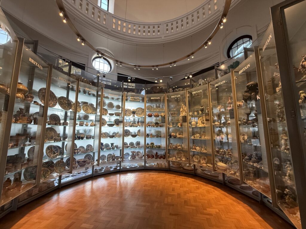 A circular museum gallery with curved glass display cases lining the entire wall, filled with a vast collection of British ceramics. The room features a herringbone wooden floor and a high, domed ceiling.