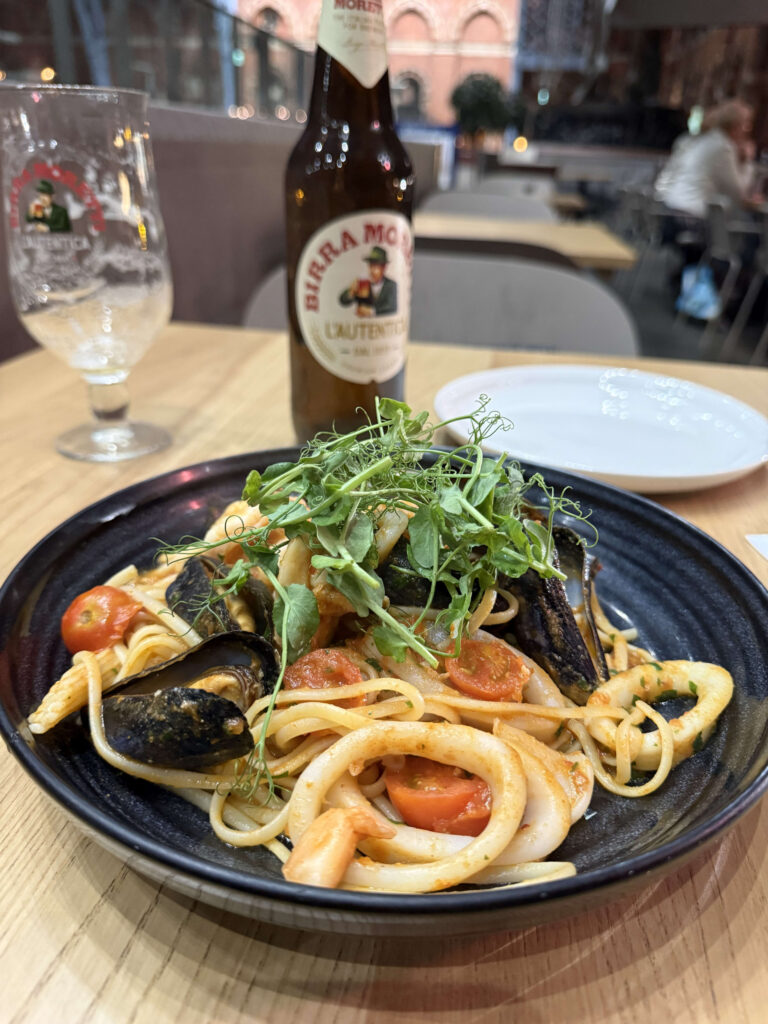 Seafood linguine topped with mussels, calamari, and tomatoes in a black bowl, with a bottle of Birra Moretti beer in the background