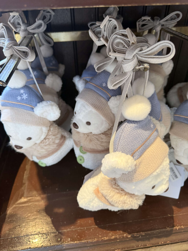 White plush bear shoulder bags, identified as Duffy, hanging on a store rack wearing blue and beige knit winter hats and faux fur capes