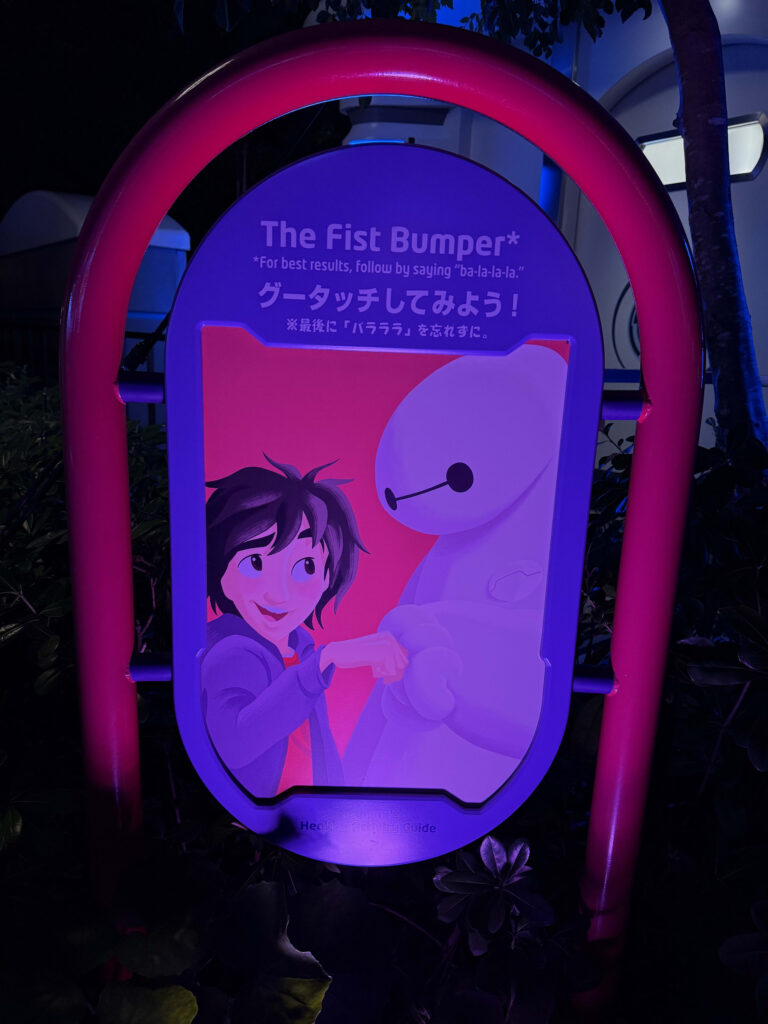 「The Fist Bumper」と書かれた紫色のライトアップされた看板で、ヒロとベイマックスがグータッチをしている様子