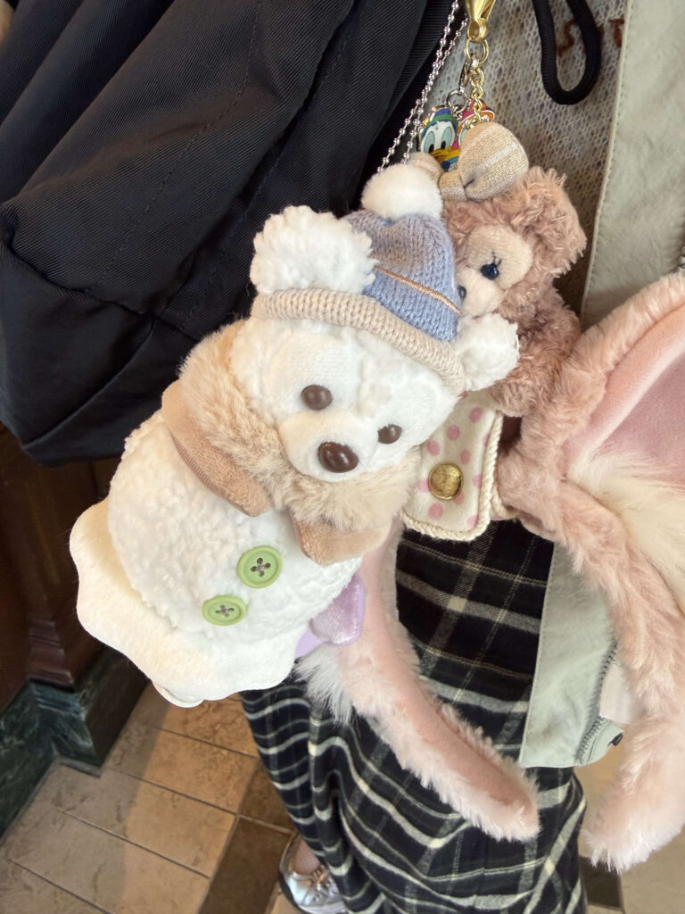 A white snowman-themed bear plush wearing a blue hat and a ShellieMay plush charm attached to a black shoulder bag strap