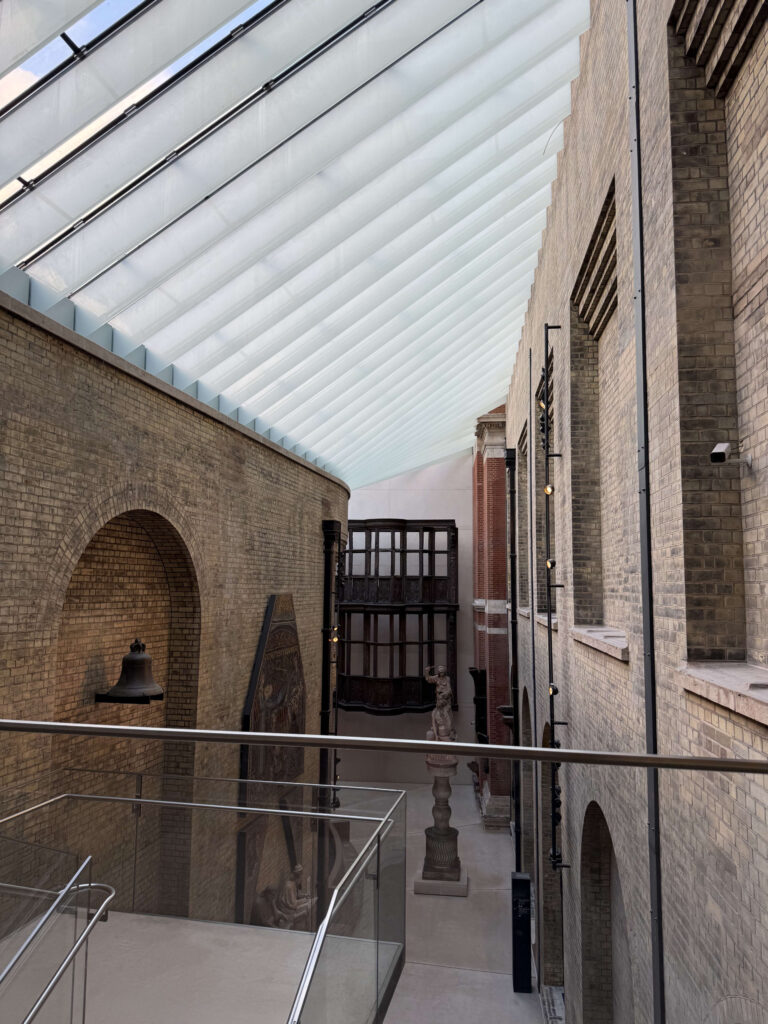 A museum corridor enclosed by brick walls with natural light streaming in from a glass roof. Art exhibits are visible on the lower level.