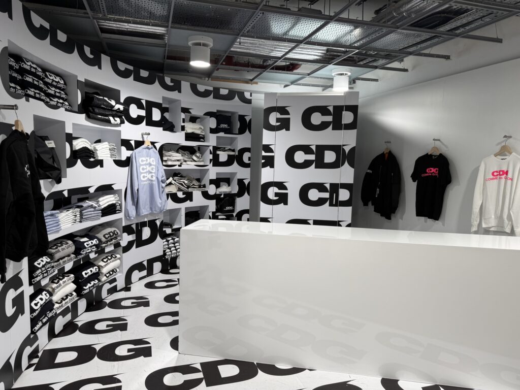 Interior of a Comme des Garçons store with walls and floors covered in black and white CDG logos, displaying clothing on shelves and racks