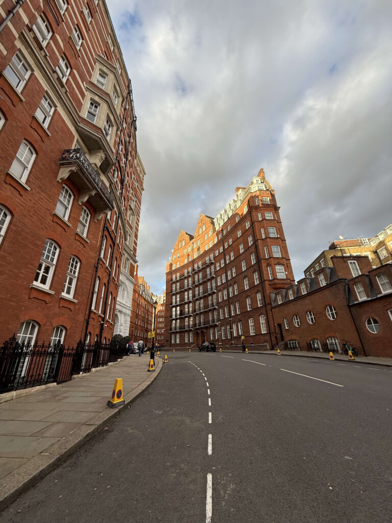 A wide-angle, low-angle view of a curving street in London, flanked by tall, elegant red-brick residential buildings under a partly cloudy sky.