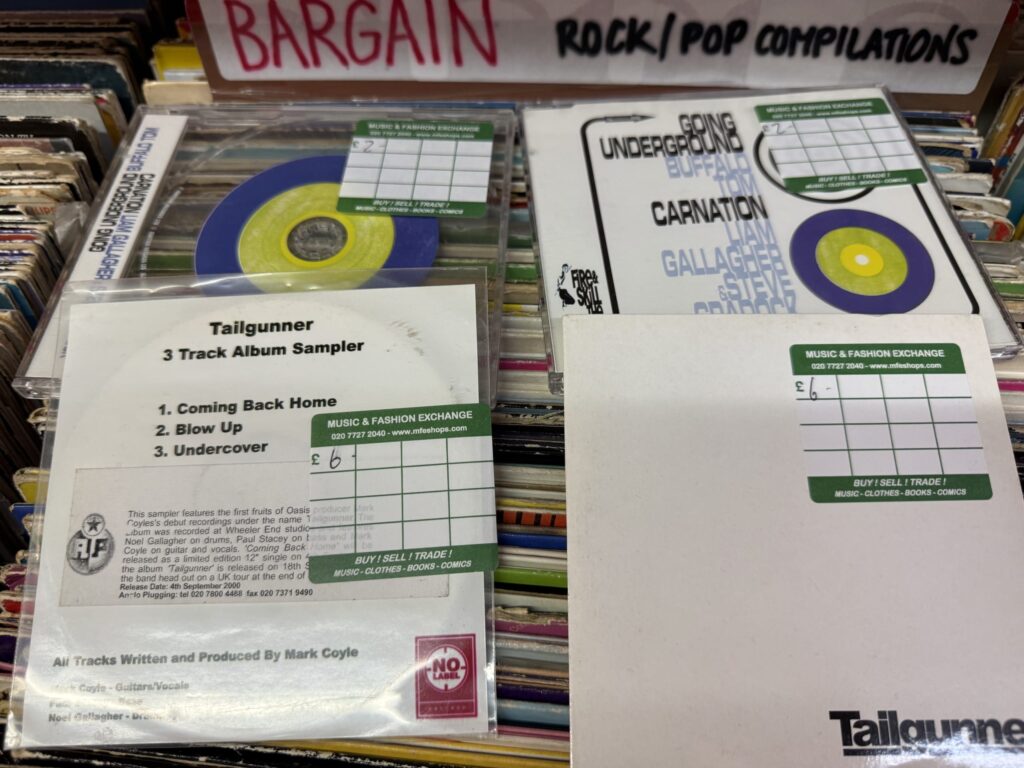 CDs in a record store bargain bin, including Liam Gallagher's 'Carnation' cover and a Tailgunner promo CD featuring Noel Gallagher