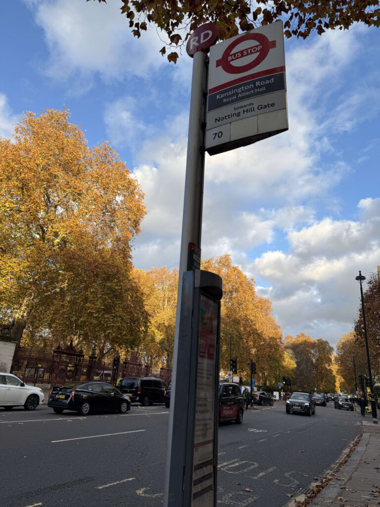 A London bus stop sign on a sunny autumn day. In the background are trees with golden leaves and a blue sky. The sign reads 'Kensington Road' and 'towards Notting Hill Gate'.