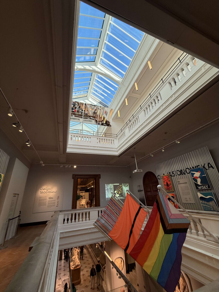 A view of a museum's multi-story atrium from an upper floor. Bright light streams in from a large skylight, and colorful flags, including a rainbow flag, hang from the railing in the foreground.