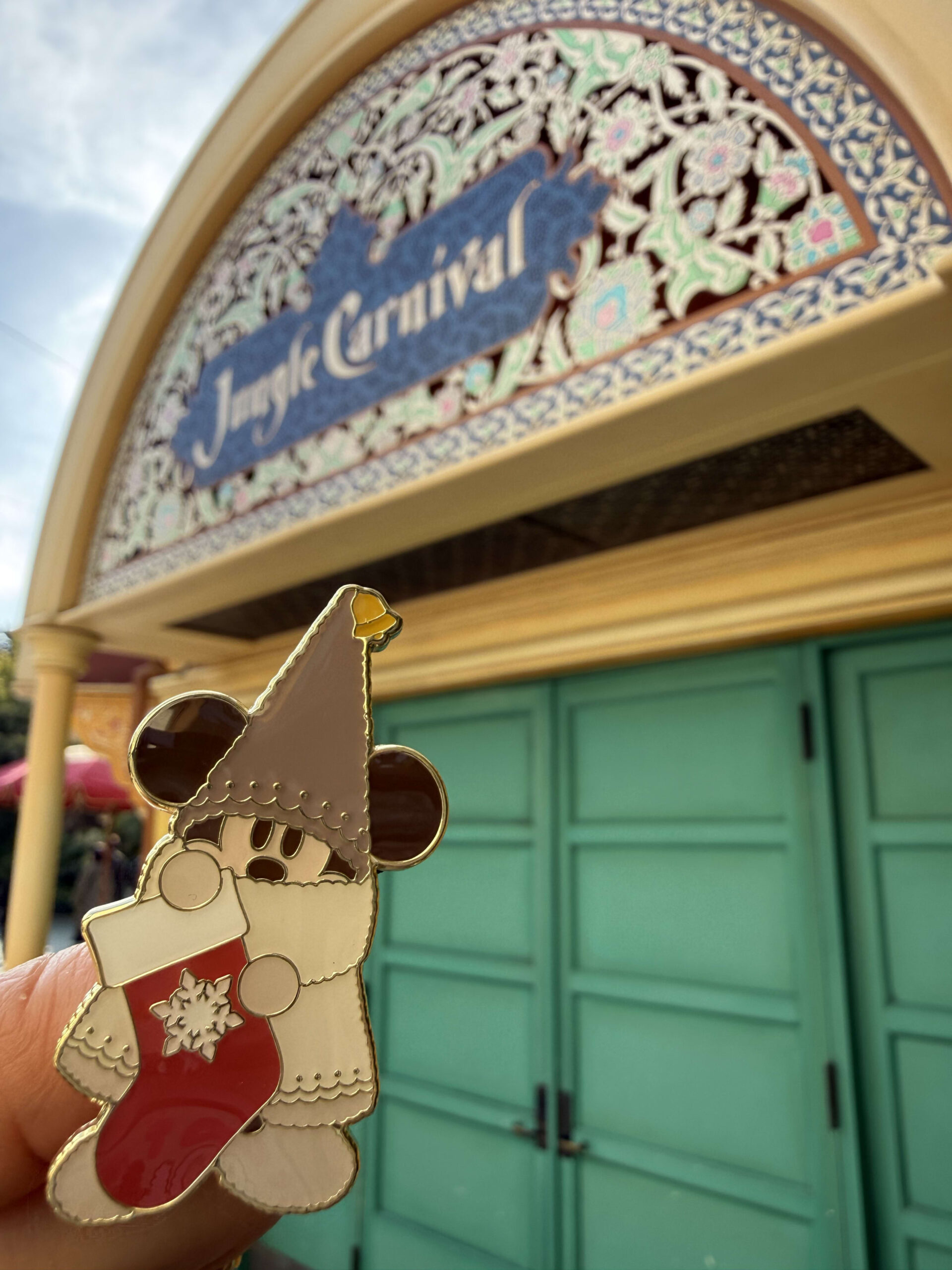 東京ディズニーランドのジャングルカーニバルの看板を背景に、手に持ったクリスマスの妖精リルリンリンのピンバッジ