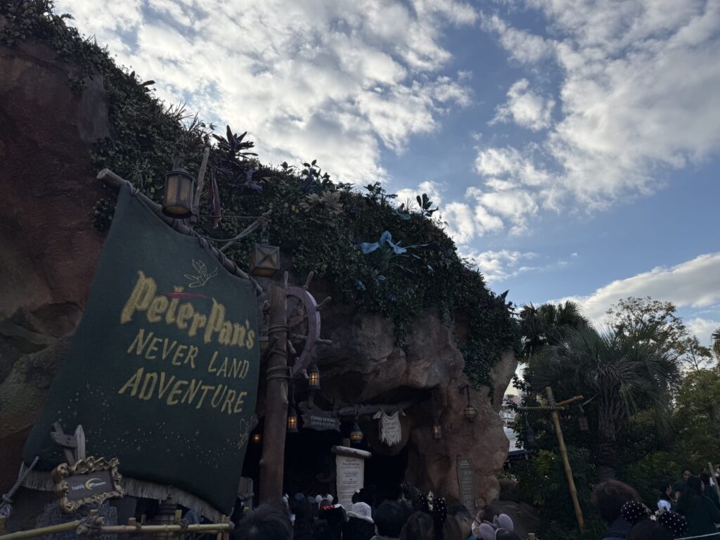 青空の下、「Peter Pan's NEVER LAND ADVENTURE」と金色の文字で書かれた緑色の帆の看板と、植物に覆われた岩山のアトラクション入り口