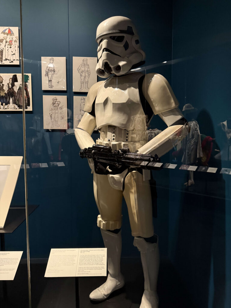 博物館の青緑色の壁を背景に、ガラスケースの中に展示されているスター・ウォーズのストームトルーパーの全身コスチューム。ブラスターライフルを手に持っている。背景にはコンセプトアートのスケッチが見える。