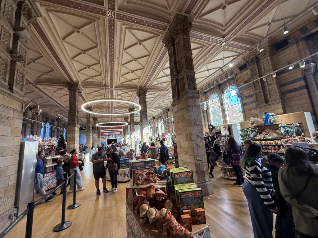 Interior of the Natural History Museum gift shop in London, featuring ornate terracotta ceilings and shelves stocked with dinosaur plush toys and souvenirs.