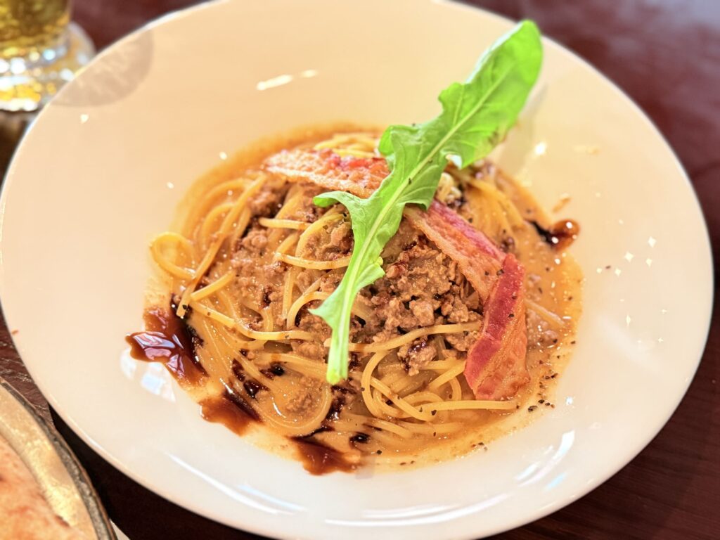 Spaghetti with beef ragu sauce served in a white bowl, topped with a slice of bacon and a green leaf