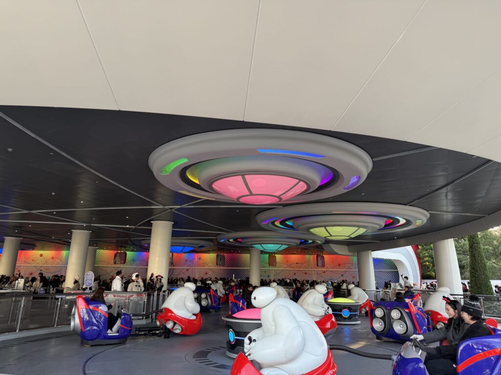 東京ディズニーランドの「ベイマックスのハッピーライド」で、カラフルな近未来的照明の下、ケア・ロボットが引っ張るライドを楽しむゲストたち