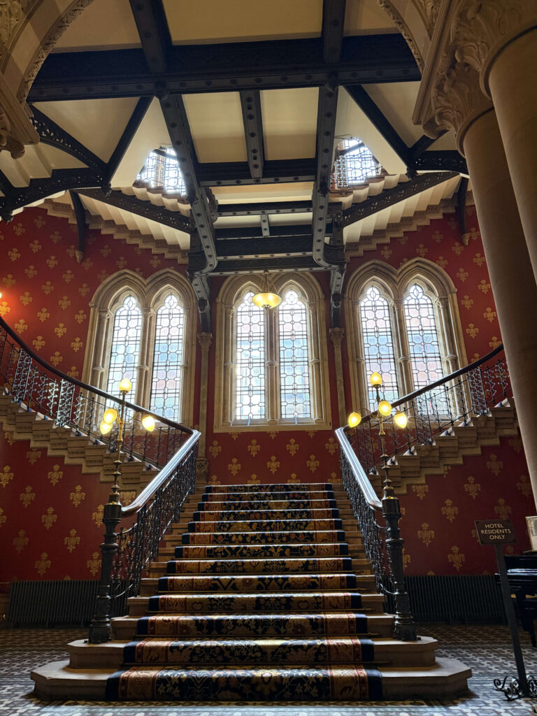The grand staircase at the St. Pancras Renaissance Hotel in London, featuring red patterned wallpaper, arched windows, and an intricate Gothic Revival ceiling