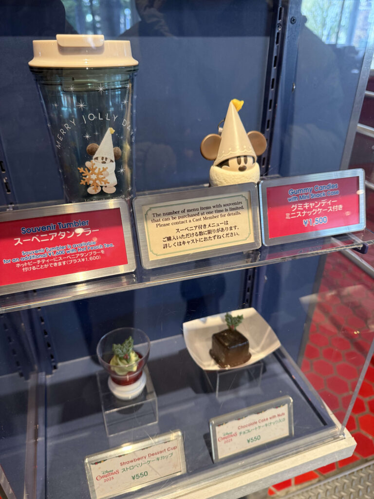 A display case featuring Disney Christmas souvenirs with Li'l RingRing designs, including a tumbler and mini snack case, along with a strawberry dessert cup and chocolate cake