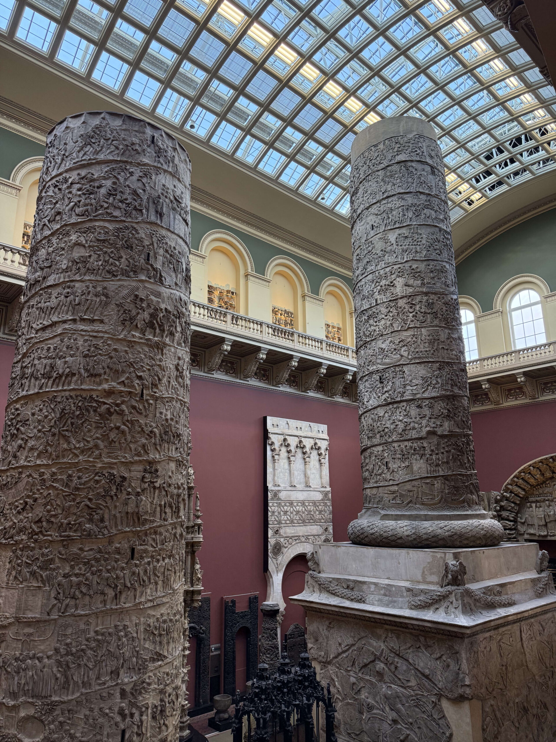 A massive replica of Trajan's Column stands in a tall museum hall. The column is covered in detailed reliefs and is illuminated by light from a glass ceiling above.