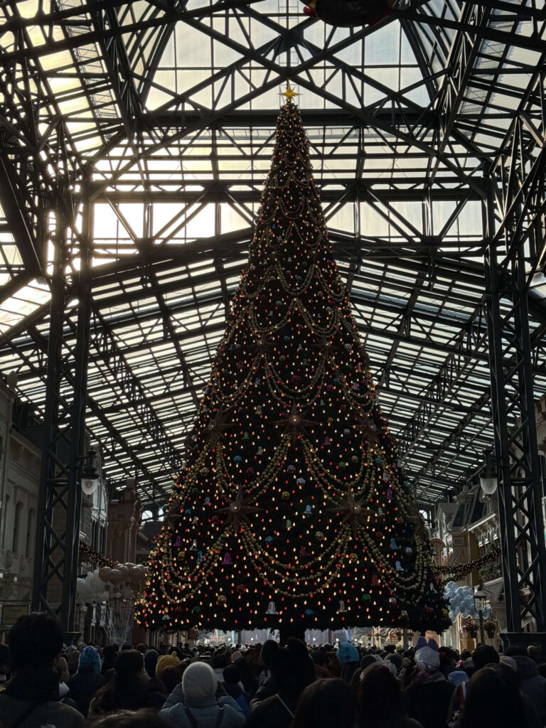 A towering Christmas tree decorated with colorful lights and golden garlands standing under a large glass-roofed atrium with a crowd of people below.