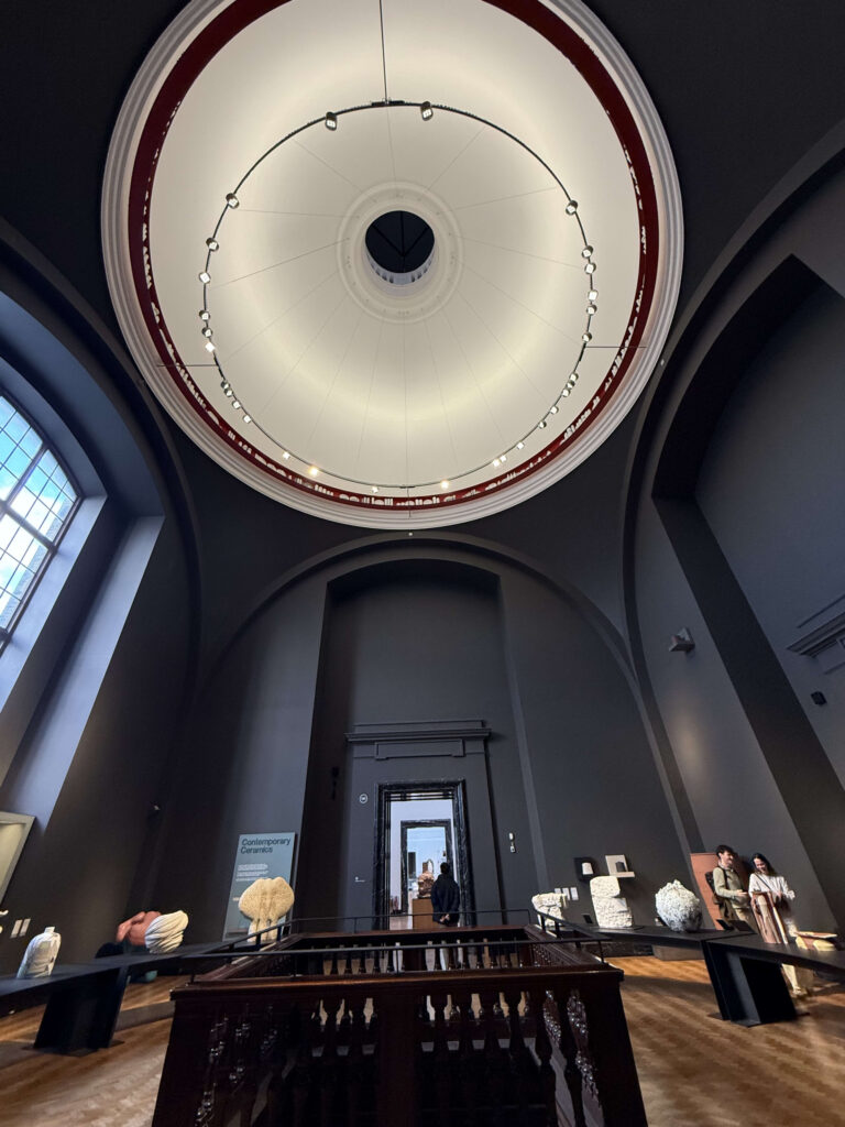 A low-angle shot looking up at a giant white dome ceiling with a beautiful circular light fixture, set against the dark walls of a museum's open hall.
