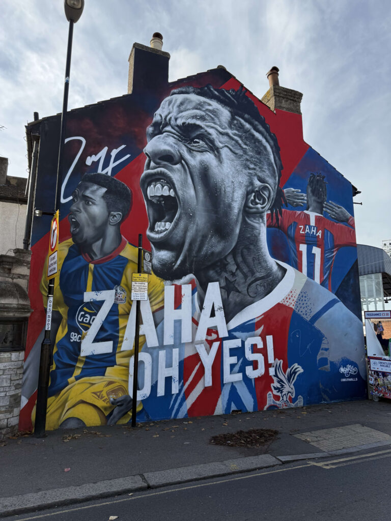 Large mural on the side of a building depicting Wilfried Zaha with a screaming expression and the text 'ZAHA OH YES!'.