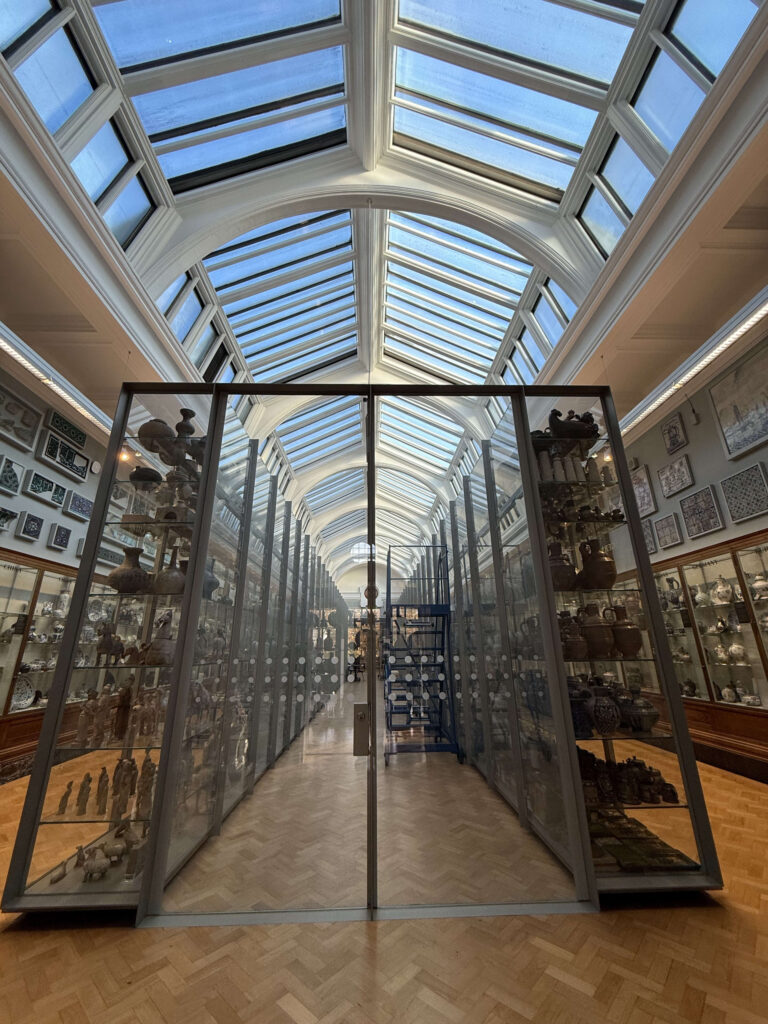 A long museum gallery with a high, glass-paned ceiling. On both sides are tall glass display cases filled with a vast collection of pottery and ceramics.