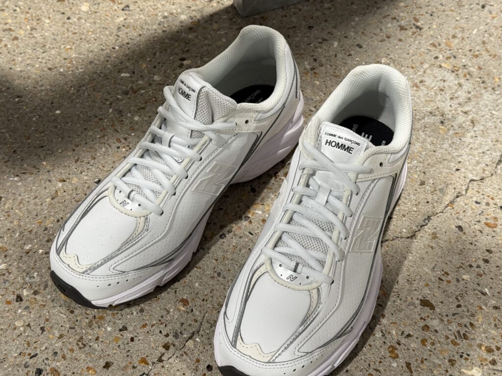 自然光の下、コンクリートの地面に置かれたNew Balance U509とCOMME des GARÇONS HOMMEの白いコラボレーションスニーカー