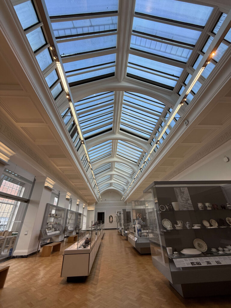 A low-angle shot looking down a long museum gallery. The prominent feature is the high, arched glass ceiling allowing natural light to fill the room. Glass display cases with ceramics line the sides, and the floor is made of wood parquet.