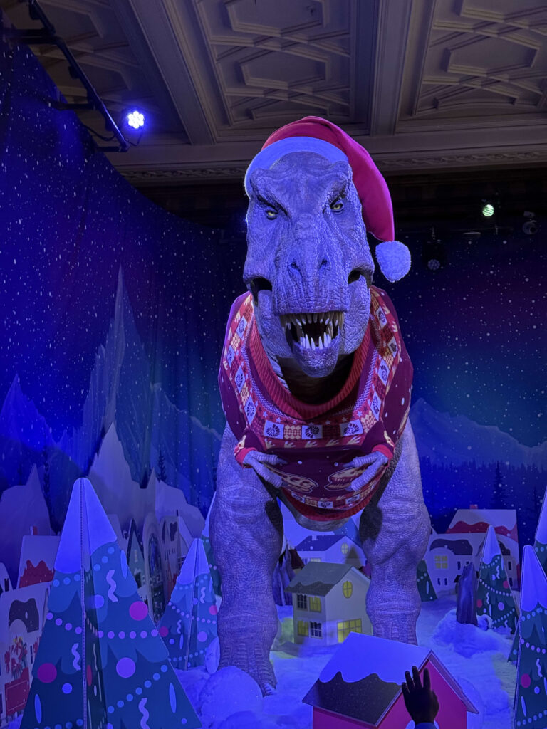 An animatronic T-Rex wearing a red Santa hat and a festive Christmas sweater against a snowy backdrop