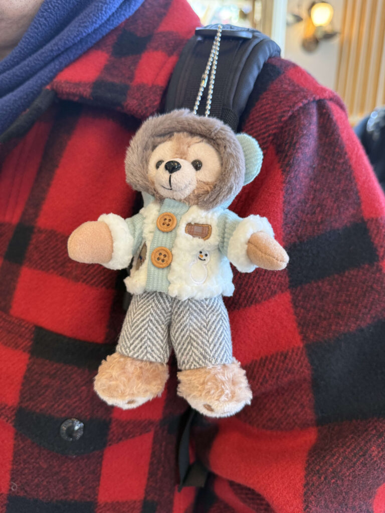 A Duffy the Disney Bear plush keychain wearing a winter coat and pants, attached to a black backpack strap against a red and black buffalo plaid shirt