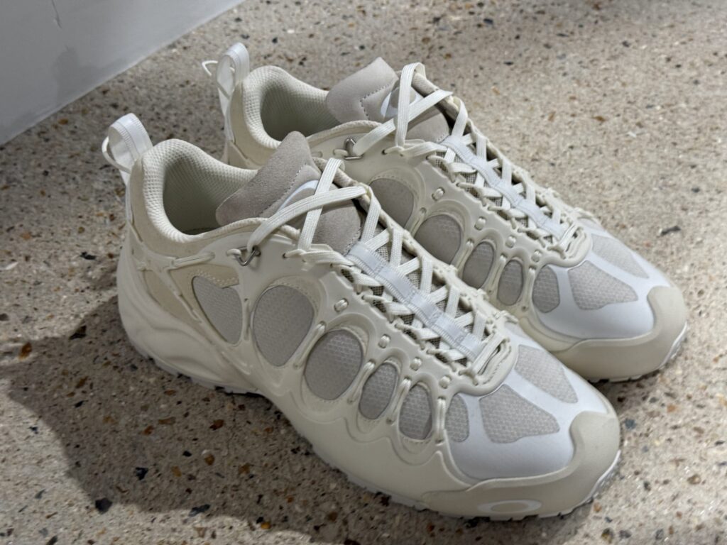 斑点模様の床に置かれたCOMME des GARCONS JUNYA WATANABE MANとOAKLEY FACTORY TEAMのコラボレーションによる白いIBEXスニーカー