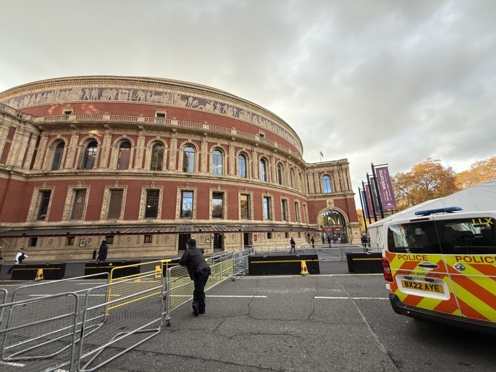 The exterior of the Royal Albert Hall in London under a cloudy sky. In the foreground, a police officer stands guard by barricades, with a police van parked to the right.