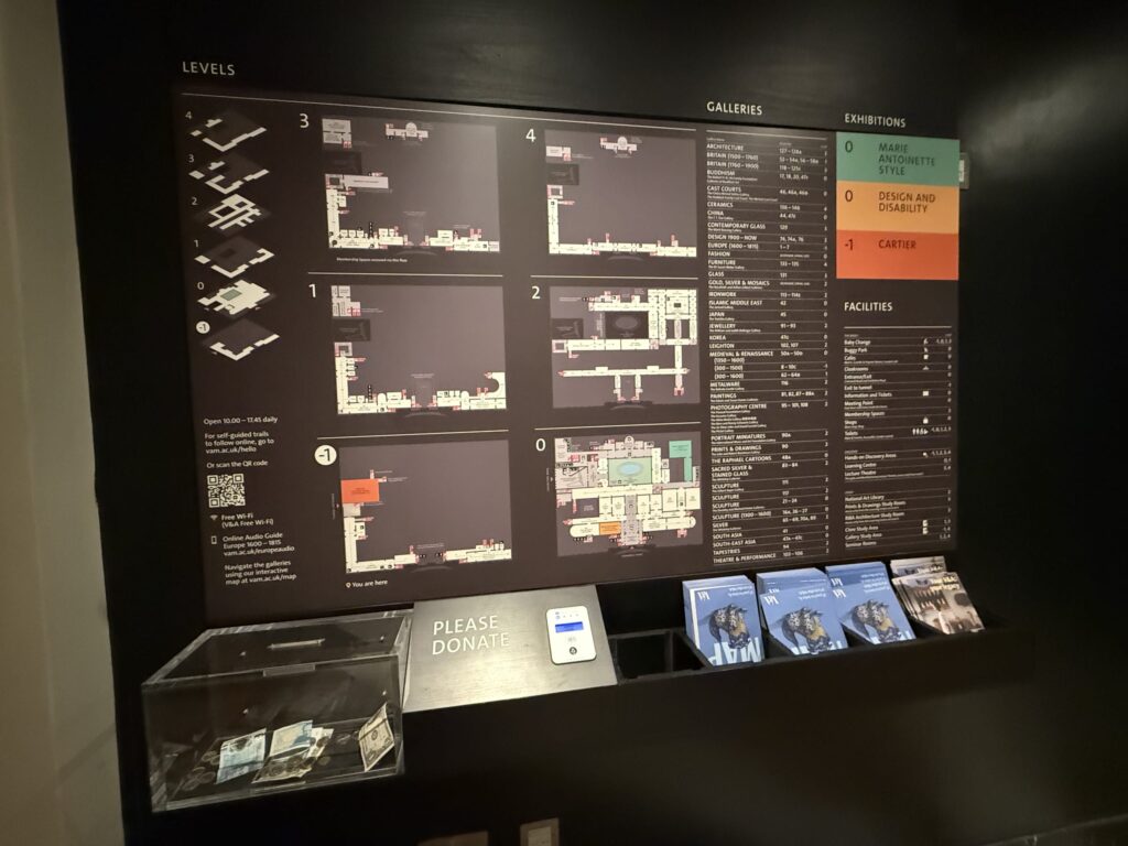 An illuminated museum guide map on a dimly lit wall. It shows detailed floor plans and exhibition information, with a donation box and leaflets placed in front of it.