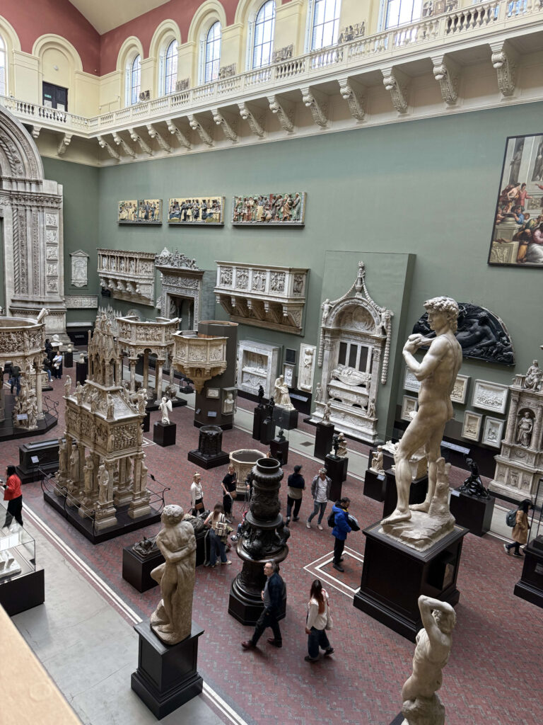 A high-angle view of a grand museum hall filled with plaster casts of ancient and Renaissance sculptures and architecture. A full-size replica of Michelangelo's David is a centerpiece, surrounded by visitors exploring the vast collection.