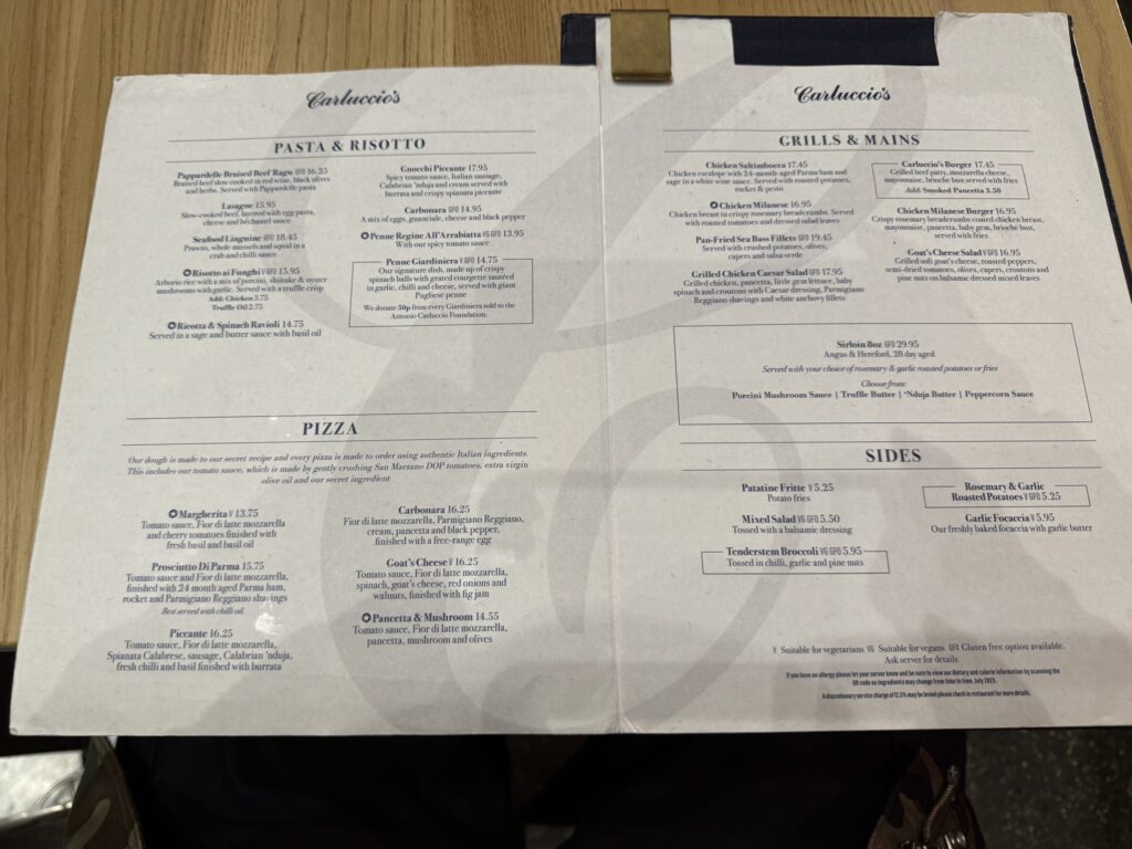 An open menu from Carluccio's restaurant resting on a wooden table, displaying sections for Pasta & Risotto, Pizza, Grills & Mains, and Sides.